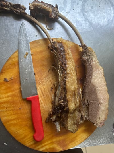 The image depicts two bone-in cuts of cooked meat on a round wooden cutting board. A large knife with a red handle lies to the left of the meat. The meat appears to be roasted or braised, with some browning and visible fat. The cutting board is placed on a metallic surface, likely a kitchen counter or table. There are no discernible elements in the image that would indicate a specific location within the city of Kostanay, Kazakhstan.
