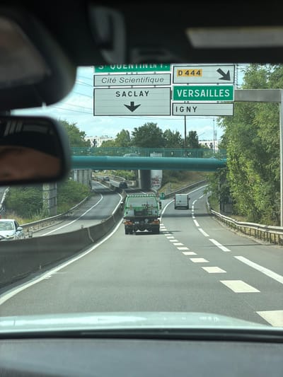 This media file captures a view from a vehicle driving on a highway in Palaiseau, France. The scene is dominated by road signs and the surrounding environment, suggesting a bright, possibly sunny day with lush green trees lining the road. The perspective is from inside the car, looking forward and slightly to the side, with part of the car's interior, including a rearview mirror reflecting a person's face, visible in the foreground.

Multiple road signs are prominent. One sign indicates "VERSAILLES" in green text with an arrow pointing to the right. Below it, another sign reads "IGNY" in white text on a green background, also with a rightward arrow. A yellow sign displays "D 444" with a black arrow pointing left. Another set of signs shows "Cité Scientifique" and "SACLAY" with rightward arrows. The road itself is asphalt, marked with white lane dividers and diamond shapes.

A green overpass spans the highway in the mid-ground. Several vehicles are visible on the road, including a white van and a green truck with a visible license plate and rear lights. The overall impression is one of a typical daytime drive on a French highway, with clear signage guiding traffic towards various destinations. The presence of trees and the bright lighting suggest it is daytime, and the weather appears to be fair.
