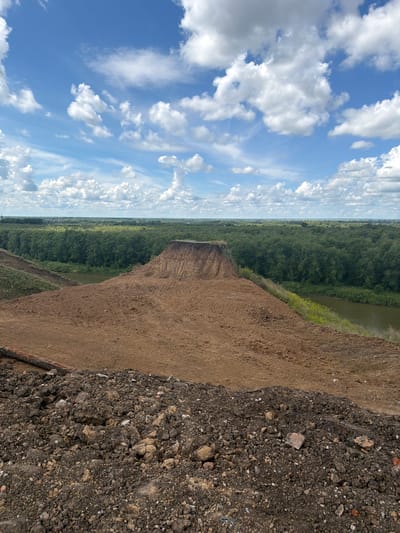 The image depicts a hilly, eroded landscape under a bright blue sky with scattered white clouds. In the foreground, a pile of dark, clumpy soil with visible rocks and debris dominates the bottom of the frame. Behind this, a large mound of light brown earth slopes downwards. Beyond the mound, a dense green forest stretches across the middle ground, bordering a calm body of water that reflects the sky. In the distance, a faint line of trees and buildings suggests a settlement or city. The overall impression is one of a natural or altered landscape, possibly a quarry or a site undergoing earthworks, located within a region with significant vegetation and water features. The location is identified as Petropavl City Administration, Kazakhstan.