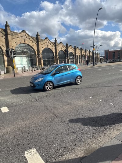 A blue Ford Fiesta is parked on the side of a road next to Sheffield Train Station. The station is a large, sandstone building with arched windows and a peaked roofline, flying a Union Jack flag. There are pedestrians walking along the pavement adjacent to the station. The sky is blue with cumulus clouds. The road is asphalt with white lane markings. A street light is visible in the background. A modern, brick building can be seen behind the station.