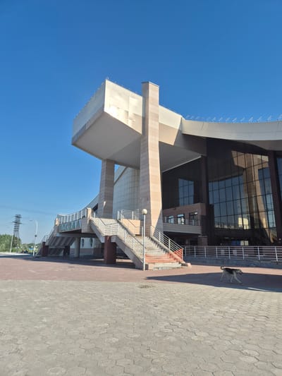A low-angle, full outdoor shot captures the modern, angular architecture of a train station under a clear blue sky in Nizhnevartovsk, Russia. The building features a prominent cantilevered section with a metallic facade, supported by beige stone pillars. A large, multi-tiered staircase with white railings leads up to the elevated structure. The lower part of the building is dominated by a dark glass facade, reflecting the sky and surroundings. A paved plaza with geometric stone tiling extends in the foreground. To the left, a distant power line tower and trees suggest an urban setting. In the middle-ground right, a medium-sized dog with a tan and white coat runs across the plaza. There are no visible people in the immediate vicinity of the building's entrance. The scene conveys a sense of calm and quiet during daylight hours.