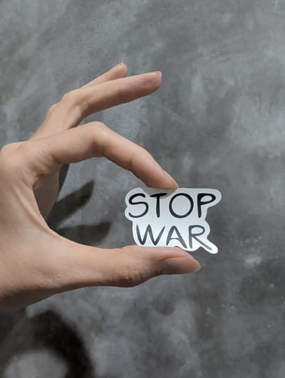 A human hand holds a white sticker with the words "STOP WAR" written in black. The background is a mottled gray surface. There is no information in the image to indicate a location in Ko Pha-ngan, Thailand.