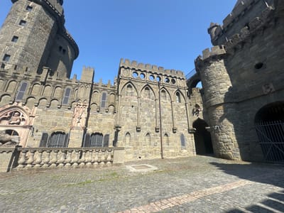 The image captures the exterior of **Löwenburg Castle**, a medieval-style castle located in **Kassel, Germany**. The castle is constructed of stone and features imposing towers, battlements, and arched windows with shutters. The foreground shows a cobblestone courtyard with a patterned design, and the sky is a clear, bright blue. The architecture is detailed and appears well-maintained. There are no people visible in the image, and the scene is static, focusing solely on the architectural elements of the castle and its surroundings.
