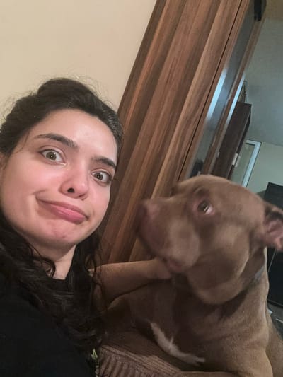 This is a close-up selfie of a young woman and a dog, taken indoors. The woman is in the foreground, looking directly at the camera with wide, surprised eyes and a slightly puckered mouth. Her dark hair frames her face. To her right and slightly behind her is a brown dog, a pitbull mix, whose face is blurred due to motion. The dog appears to be looking towards the woman.
The background suggests an indoor environment with wooden paneling and a hint of furniture. The lighting appears to be artificial, suggesting it could be evening or night. The overall mood is playful and intimate, capturing a candid moment between the woman and her pet. There is no visible text in the image.