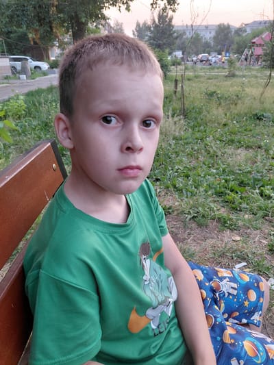 The media file shows a close-up, vertically oriented shot of a young boy sitting on a wooden bench outdoors. He is wearing a green t-shirt with a cartoon character print on it and blue shorts with a space-themed pattern featuring astronauts and planets. The boy is looking towards the camera with a neutral expression.
In the background, there is a grassy area with some scattered leaves, and beyond that, a playground with some structures and parked cars are visible. Trees and buildings are also present in the distance, suggesting an urban or suburban park setting. The lighting suggests it is either early morning or late afternoon. The weather appears to be mild and clear.
No visible text is present in the media file. The scene is likely a casual outing at a park in Oskemen, Kazakhstan.