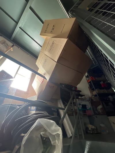The image, taken from a low angle looking upwards, depicts a densely packed storage area, likely a warehouse or backroom in Campana, Argentina. The scene is dominated by numerous cardboard boxes stacked on and around metal shelving units and support beams. Several boxes bear the label "FRAGIL" and the brand "emilux," with text indicating "INDUSTRIA ARGENTINA" on multiple surfaces.
In the background, what appear to be musical instruments, specifically the necks and bodies of guitars, are visible amongst other stored items on shelves. Further back, electronic appliances or devices are stacked, one clearly showing the brand "PHILCO." To the right, a large coiled object, possibly a roll of industrial cable or tubing, is stored next to a white plastic bag.
Bright natural light streams in from a window, suggesting it is daytime. The overall impression is one of significant clutter and organized, albeit dense, storage. Other visible text includes "Indelable," "mantia de buen are," "01," "Ind," and "TUT."