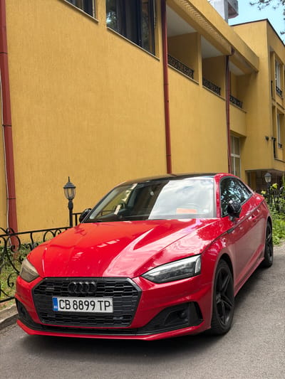 A red 2020-2024 Audi A5 Coupe is centrally parked on an asphalt surface, angled slightly towards the right. The vehicle features a gloss black roof, black multi-spoke alloy wheels, and a black honeycomb grille displaying the Audi rings logo and a "Quattro" badge. A Bulgarian license plate, "BG CB 8899 TP," is affixed to the front bumper. The car's body reflects bright daylight, showing sky and surrounding structures.
Behind the vehicle stands a multi-story building with a textured, mustard-yellow stucco facade. Red vertical downspouts or pipes are visible on the building's exterior, notably on the left and near the center. The building incorporates recessed balconies on its upper floors, each featuring ornate black wrought iron railings. Dark-framed windows are present across different levels, some exhibiting reflections.
To the left of the Audi, a black wrought iron fence with decorative elements separates the pavement from a small area of green foliage. A black vintage-style street lamp with a glass lantern is positioned behind this fence, near the vehicle's hood. Another similar lamp is partially visible on the right side of the image, behind the car's rear quarter panel, amidst green plants. The pavement is dark gray asphalt. No individuals are depicted in the scene. The image is brightly lit, suggesting daytime conditions. The setting is identified as the city of Kyustendil, Bulgaria.