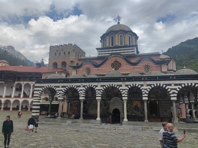 The image captures the bustling courtyard of Rila Monastery in Bulgaria under a cloudy sky. The central focus is the magnificent main church, characterized by its distinctive architecture. Its lower facade features striking horizontal red and white stripes, topped by a large central dome adorned with vertical yellow and dark stripes, surmounted by an Orthodox cross. Numerous smaller domes are also visible.
A prominent feature of the church is its arcaded narthex, where columns support arches with alternating black and white stripes, creating a visually impactful pattern. Beneath these arches, intricate and colorful murals depicting religious scenes cover the exterior walls, adding rich detail to the structure.
To the left of the main church, a robust stone tower stands with crenellated battlements and arched openings, suggesting a historical defensive purpose. Other monastery buildings with red tiled roofs and white arcades are visible to the left, forming the perimeter of the paved courtyard.
The courtyard itself is made of irregular stone blocks. Several people are present, indicating a popular tourist destination. In the foreground, a woman in dark sportswear walks towards the left, while nearby, a man in white shorts carries red bags. On the right, two men are seen, one holding a smartphone to take a selfie. Other visitors can be seen walking or sitting further within the complex.
In the background, lush, forested mountains rise, with glimpses of snow-capped peaks under the overcast sky, highlighting the monastery's secluded and scenic location. The time of day appears to be daytime, with soft, diffused light. No clearly legible text or specific dates are prominent on the buildings, though some faint markings on the main dome are barely discernible. The overall atmosphere is one of serene historical grandeur mixed with active tourism.
