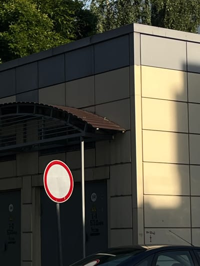 The image shows a segment of a modern building with a dark grey and light beige facade, possibly a commercial or industrial structure, with a dark-colored awning over an entrance area. In the foreground, a circular traffic sign with a red border and a white center, indicating a prohibition or restriction, is mounted on a pole. To the left and right of the sign are doors or panels with markings that appear to be electrical or utility access points, featuring labels such as "T-2 530" and "T-1 530". Trees with lush green foliage are visible in the upper left corner. A shadow is cast across the right side of the building. In the lower part of the image, the roof and hood of a dark-colored car are visible. The scene appears to be outdoors, likely in an urban setting. Without specific landmarks or context, the precise location in Vitsebsk, Belarus, cannot be determined from the visual information alone.