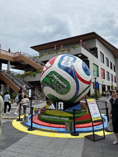In Qingpu District, China, a large replica of a FIFA World Cup soccer ball is displayed outdoors in front of a modern building complex. The soccer ball is colorful, with "TRIONDA" and "adidas outlet" branding visible. It is situated on a circular base decorated with the words "ARGENTINA," "BRAZIL," and "DEUTSCHLAND" in large letters on concentric rings. People are present in the scene, with some walking and others standing near escalators that lead up to the building. A woman in a black dress and sunglasses is in the foreground on the right, carrying a shopping bag. A sign with a QR code and Chinese text is also displayed near the soccer ball. The sky is overcast, and the overall atmosphere suggests a commercial or entertainment area.