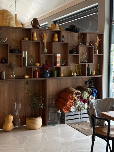 The image shows a wooden shelving unit filled with various decorative items. The shelves are organized into cubbies of different sizes. On the top shelf, there are two woven lampshades, a brown jug, and a straw hat. Below these, on the second shelf, are several gourds, a teapot, and some dried flowers. The third shelf holds more decorative bottles, a blue vase with red berries, and a green bottle. On the fourth shelf, there are various glass bottles, a small plant in a woven pot, and a few dried gourds. The bottom of the shelving unit is partially visible, and it appears to be a wall. In front of the shelving unit, there are stacked colorful blankets and an umbrella. A chair with a grey garment draped over it is positioned at a wooden table on the right side of the image. The floor is tiled with a light-colored, patterned tile. The overall impression is one of a curated display of rustic and decorative items. The image is taken indoors, likely in a cafe or home environment. There are no people visible in the image. The location in Dsegh, Armenia is not discernible from the visual information provided.