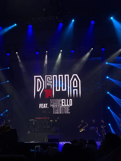 A low-angle view from a concert in Pagedangan, Indonesia, showing a stage bathed in blue and white spotlights. On a large screen, white lettering reads "DEWA 19 FEAT. MARCELLO TAHOE." A few people in dark clothing are seen on stage, some playing instruments such as a saxophone. Silhouettes of audience members can be seen in the foreground.
