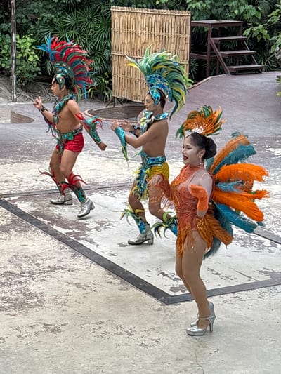 A group of three performers in elaborate feathered costumes are dancing on a paved outdoor stage. The performer in the foreground, a woman, wears a bright orange, sequined leotard with large orange and turquoise feather wings and a feathered headdress. She is wearing silver high-heeled shoes and orange gloves. To her left, a male performer wears a red sequined loincloth, a blue and green feathered headdress, and silver boots adorned with red feathers. Behind him, another male performer also wears a colorful feathered headdress and costume. The stage is bordered by dark lines and has a textured concrete surface. In the background, there is a woven bamboo screen and lush green foliage, suggesting an outdoor setting in a tropical location. A wooden step stool is visible to the right of the bamboo screen. The lighting suggests it is daytime.