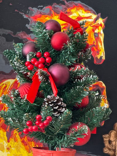 A close-up shot features a Christmas wreath with a backdrop that appears to be a painting of a horse in flames. The wreath is composed of artificial evergreen branches, dusted with white snow-like material. It is adorned with several red and deep purple ornaments. Sprigs of red berries and a pine cone are also incorporated into the wreath. Red ribbons are tied in bows around some of the branches.
The background painting depicts a horse, rendered in abstract, fiery colors of yellow, orange, and red, against a dark background. To the right of the wreath, a portion of a golden, ornate object is visible, possibly a statue or decorative item. The overall impression is a festive and somewhat dramatic composition, blending traditional Christmas elements with a bold artistic statement. No specific time of day, weather, or location cues are discernible, beyond the user-provided context of Tambov, Russia. There is no visible text in the image.