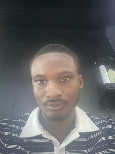 A close-up portrait of a dark-skinned man wearing a striped shirt with a white collar. The background is dark and indistinct, partially revealing what appears to be the interior of a vehicle. The location is Lugbe, Nigeria.