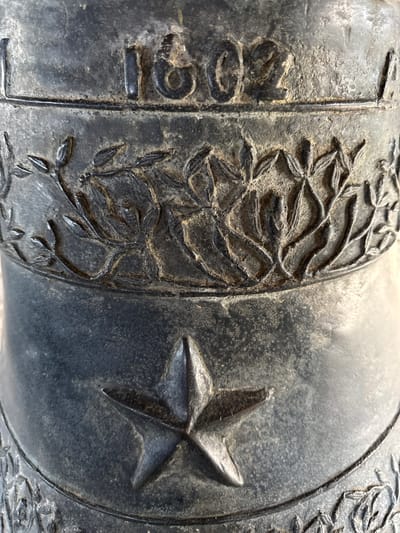 The image shows a portion of an aged, dark-colored cannon, likely made of metal. The number "1692" is inscribed vertically on the left side. A band with a detailed, raised design of laurel or olive branches encircles the cannon. To the right of the branches, a prominent five-pointed star is embossed. The background is out of focus, indicating that the camera was focused on the cannon details. The location of this image is Gerokgak, Indonesia, as specified in the prompt.