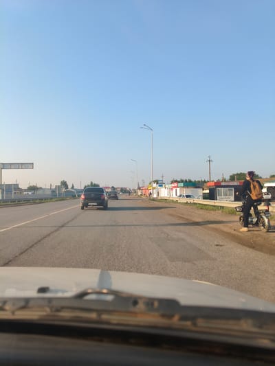 The image depicts a clear sky over a two-lane asphalt road in Подстепной ауылдық округі, Kazakhstan. The road appears to be a highway or a main thoroughfare, with a grassy shoulder on the right side lined with several small, colorful kiosks or shops. A modern black sedan is driving in the right lane, and a dark-colored car is visible further ahead in the same lane. To the right, a young person wearing a dark top, black pants, and a purple headscarf is standing next to a small electric scooter. They have a beige backpack slung over their right shoulder. The scooter is parked on the side of the road, and the person is facing away from the camera. The kiosks on the right feature various signs, some with Cyrillic script. Streetlights are visible on both sides of the road, and power lines run above the kiosks. The foreground shows the blurry edge of a vehicle's hood and windshield wipers, suggesting the image was taken from inside a car. The overall scene is bright and bathed in natural light, indicating it is daytime, likely late afternoon or early evening due to the warm tones.