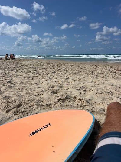 The image depicts a beach scene at Beit Yanai, Israel, viewed from a low perspective on the sand. In the immediate foreground, the right leg of an individual, covered in brown hair and wearing blue swim trunks with a white stripe, is visible alongside an orange surfboard. The surfboard, positioned diagonally, features the black text "MULLET" with a flame graphic and has a blue rail. The sand in the foreground and midground is light brown, showing various textures and indentations.
In the midground, a sandy beach extends towards the water. To the far left, two individuals are seated on what appear to be beach chairs. Closer to the waterline, several people are partially submerged in the turquoise-blue Mediterranean Sea, which exhibits moderate waves with white crests. The background consists of a clear blue sky dotted with scattered white cumulus clouds. The lighting suggests a bright, sunny day.