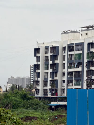 The image depicts a dense urban landscape in Vasai-Virar, India, characterized by multi-story residential buildings and lush vegetation. The foreground is dominated by a thick growth of green bushes and plants, with some patches of soil visible. In the mid-ground, a tall, white apartment building with dark blue accents stands prominently, featuring numerous balconies and windows. Some balconies display laundry hanging out to dry, and potted plants. Behind this building, several other residential structures of varying heights and architectural styles are visible, including a taller, older building with a repetitive, grid-like facade. The sky is overcast, casting a diffused light over the scene. A temporary blue metal barrier is positioned in the right foreground, partially obscuring the lower portion of the apartment building. The overall impression is one of a developing urban area with a mix of modern and older structures, interspersed with pockets of greenery.