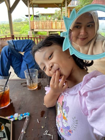 At a table in Tamiajeng, Indonesia, a girl in a pink Daisy Duck sweatshirt sits beside a woman reflected in a blue-framed mirror. The girl touches her mouth. On the table are two glasses of iced tea, a beaded phone strap, and scraps of food. In the background are rice fields seen through the open structure of a building. The girl wears denim.