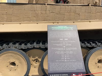 The image shows a close-up of the tracks and road wheels of a German Panzerkampfwagen VI Ausf. E Tiger heavy tank, a museum exhibit at the Military Technology Museum in Verkhnyaya Pyshma, Russia. A descriptive plaque, with text in Russian and English, is positioned in front of the tank. The plaque details the tank's specifications and history, noting it was developed in 1942 and saw action on all fronts of World War II. The tank's camouflage exterior is visible, with a textured pattern and a thick rope running along the top. The environment appears to be outdoors, with sunlight casting shadows. No people are visible in the frame.