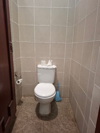 The image shows a bathroom located in a building at latitude -0.772, longitude 36.421 in Karagita, Kenya. The bathroom is tiled in a light beige color with a white toilet situated in the corner, facing the wall. There are two rolls of toilet paper on the wall behind the toilet and a toilet brush on the floor. The toilet lid is down and the toilet seat is up. The floor is tiled in a darker beige color. There is a brown wooden door frame in the lower right corner.
