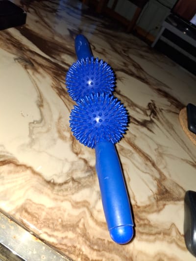 A blue massage roller with two spiked balls is placed diagonally on a marbled surface. The roller is positioned with the spiked balls towards the top left of the image and the handle extending towards the bottom right. The background is blurred, showing furniture and a hint of a window. The lighting suggests an indoor environment. The specific location in Åsgårdstrand, Norway, is not discernible from the image.