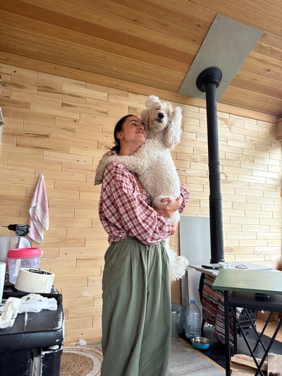 A young woman with dark hair, wearing a red and white plaid shirt and olive green wide-leg pants, is holding a fluffy white poodle dog in her arms. The woman is smiling and looking upwards. The dog is being held in a playful manner, with its front paws raised. They are standing indoors, in a room with walls and ceiling paneled with light-colored wood. A black metal stove pipe extends from a gray metal plate on the ceiling, down to the floor. To the left, a pink and white striped towel hangs on the wall, and a cordless drill rests on a black surface with some white paper. To the right, a large clear plastic water bottle and a blue dog bowl are on the floor. A multi-colored woven rug is visible on the floor. The scene appears to be rustic and domestic.