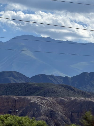 The image shows a mountainous landscape with layered mountains receding into the distance. The foreground features a power transmission tower and rocky, eroded terrain, with blurred green foliage at the bottom edge. The middle ground consists of dark, textured hills covered in scrub vegetation, while the background is dominated by hazy, blue-hued mountains under a cloudy sky. The overall impression is one of a vast, rugged natural environment, possibly in a semi-arid region, with man-made infrastructure present. The scene appears to be captured from a moving vehicle, suggested by the motion blur in the foreground. The location is identified as Unknown, Kyrgyzstan.