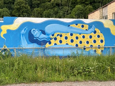 This media file is a photograph of a mural painted on a concrete wall. The mural depicts a stylized representation of a person, rendered in shades of blue and yellow. The figure appears to be floating or reclining within a cosmic or dreamlike scene.

The main subject is a human figure, likely female, with long, dark hair flowing around her head. Her body is elongated and painted in a light blue hue, contrasting with the darker blue of her hair. She is positioned on her back, with one arm raised upwards and the other bent, holding a round object that is also blue. Her eyes are closed, and her expression conveys a sense of tranquility or serenity.

Behind the figure, the mural features a bright yellow circular element adorned with a pattern of dark blue circles and small star-like shapes. This element could represent the sun, a celestial body, or a symbolic representation of energy or the universe. Wavy lines in yellow and darker blue emanate from the top of the mural, suggesting movement, light, or cosmic forces.

The mural is set against a backdrop of lush green trees, indicating an outdoor setting. The foreground of the photograph shows a strip of dense green grass and weeds, with a metal fence running horizontally across the middle of the image. Beyond the mural, a building with beige, textured walls and windows is visible, as is a portion of the sky, which appears overcast or partly cloudy.

The overall mood of the mural is peaceful and ethereal. The artwork appears to be located in a public space, possibly an urban or semi-urban environment, given the presence of the concrete wall and the adjacent building. The time of day is likely daytime, due to the natural lighting. There is no visible text in the image. The location context provided places this scene in Dilijan, Armenia.