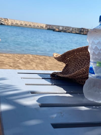 The image depicts a close-up view of a sunlit beach scene in Panormos, Greece. In the foreground, a light blue plastic lounger is visible, with shadows from palm fronds cast across its surface. Resting on the lounger is a woven straw hat. To the right, a partially visible plastic water bottle with condensation is placed. The background shows the sandy beach, the tranquil blue sea, and a distant breakwater with structures. The overall scene conveys a sense of relaxation and summer vacation.
