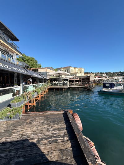 The image depicts a waterside dining establishment in Üsküdar, Turkey, likely on the Bosphorus strait, given the characteristic Ottoman-style architecture in the background. In the foreground, a weathered wooden pier with several planters containing green foliage leads towards the water. The pier is attached to a modern building with large windows, housing a restaurant called "DENİZ YILDIZI RESTAURANT," as indicated by signage. Several patrons are seated at tables both inside and on an outdoor deck area of the restaurant.
Adjacent to the restaurant, more dining spaces are visible, some with white umbrellas providing shade. The water is a deep teal color, with gentle ripples on its surface. A white and green boat is moored to the right side of the frame. Beyond the immediate structures, a hillside is dotted with residential buildings and trees. The sky is a clear, vibrant blue, suggesting a sunny day. The overall scene conveys a relaxed atmosphere of a waterfront cafe or restaurant.
