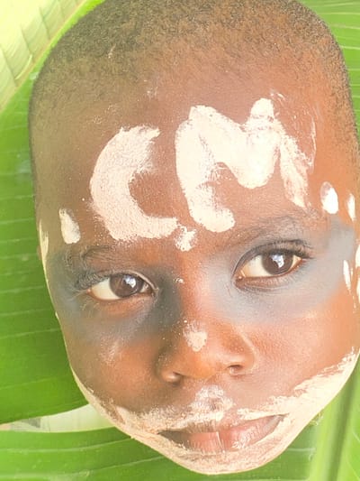 A close-up photograph depicts the face of a dark-skinned individual, possibly a child, with white markings or paint applied to the face in curved shapes. The subject is positioned on a large green leaf, which may be a banana leaf. The background is out of focus. Given the location of Achara, Nigeria, the scene might depict a cultural ritual or body adornment common in the region.