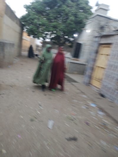 A blurred snapshot presents two women dressed in flowing garments, one green and the other red, walking along a dirt path flanked by buildings. Another figure, in a dark robe, trails behind. The scene appears to be in a village or settlement with a mix of aged concrete structures and a large tree. The ground is littered with debris, contributing to the overall sense of motion and blur.
