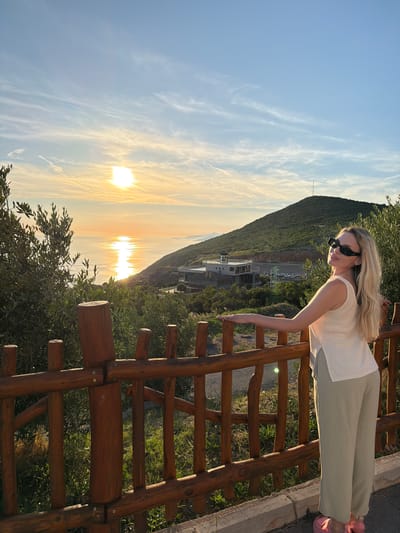 In Krimovice, Montenegro, a young woman stands by a wooden fence, looking towards a picturesque sunset over the Adriatic Sea. The sun, a bright orb reflecting on the water, casts a warm golden glow across the scene. The woman, with blonde hair and sunglasses, wears a light-colored sleeveless top and loose-fitting green pants. Behind her, a verdant hillside rises, dotted with trees and a building with a tower. The fence, made of rough-hewn logs, separates the woman from the expansive view. The sky is a soft blue, streaked with wispy white clouds, enhancing the serene atmosphere of the coastal landscape.