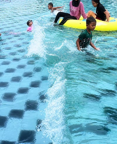 In Eampat Kwalamencirim, Indonesia, a group of people are enjoying a swimming pool. A young boy in a green t-shirt is splashing water as he walks. Further back, a child in a pink swimsuit and goggles is looking upwards, with a splash of water in front of them. To the right, two women and a child are on a yellow inflatable ring. One woman wears a pink long-sleeved shirt and a hijab, while the other is in a black t-shirt. The pool bottom is tiled with a checkered pattern of blue and dark squares. The water is clear and blue, with visible waves and splashes.