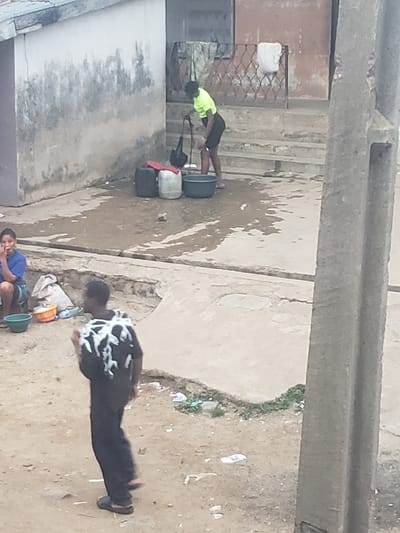 The image shows a street scene in Alalubosa, Nigeria. Three people are visible. One person wearing a yellow shirt is bending over containers. A second person in a black and white shirt is walking on the ground. A third person in a blue shirt is squatting by a bucket. The environment includes buildings, concrete, dirt, water, and various objects.