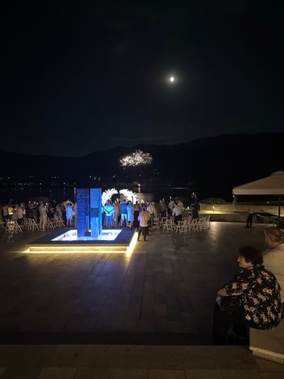 This image captures a nighttime scene in Razliv, Bulgaria. A group of people are gathered outdoors, seemingly for an event. The setting appears to be a venue with a paved area and rows of white chairs set up, suggesting a ceremony or performance. In the background, there is a body of water, possibly a lake, with distant lights indicating a town or settlement. The sky is dark, with a prominent full moon and a display of fireworks adding to the festive atmosphere.
Several individuals are visible in the foreground and middle ground, some standing and observing, while others are seated. One woman in a floral-patterned top is looking at her phone. Another man with gray hair is also visible. The lighting is a mix of ambient moonlight, stage lighting illuminating a central structure with blue and white elements, and the bright bursts of the fireworks. The overall mood seems celebratory and engaged, with people focused on the events unfolding before them. The presence of a large structure with intricate details, bathed in blue light, hints at a focal point for the gathering, possibly an artistic installation or stage. The time of day is clearly night, and the weather appears clear to allow for the visibility of the moon and fireworks.