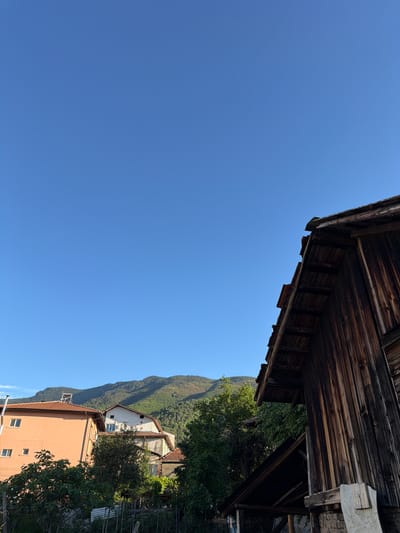 The image shows a view in Sapareva Banya, Bulgaria, with a predominantly clear blue sky. In the upper part of the frame is the corner of a weathered wooden structure, possibly a roof or wall. Below that, a landscape featuring a mountain range covered in trees is visible, along with buildings nestled in the foliage. One building is peach-colored with windows, and another is white with a red roof. Trees partially obscure the buildings.
