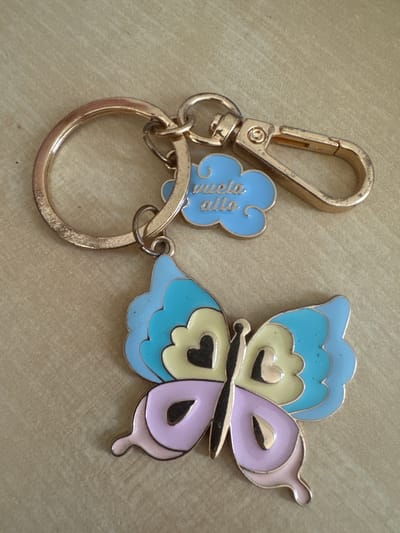 The image shows a keychain with a butterfly charm. The butterfly is made of metal and enamel, with wings in pastel shades of blue, yellow, and pink. The body of the butterfly is black. There are also two smaller charms attached to the keychain. One is a blue cloud shape with the text "vuela alto" written in cursive on it. The other charm is a simple gold-colored ring. The keychain is resting on a textured, light brown surface, possibly a table or countertop. The lighting appears to be natural and diffused, suggesting an indoor setting during the daytime. The image is a close-up shot, focusing on the details of the keychain. There are no people or other objects visible in the frame. The overall impression is that of a decorative accessory, possibly a gift. The text "vuela alto" translates to "fly high" in Spanish, which might be a motivational message associated with the keychain. Given the location context of Murcia, Spain, this could be a souvenir or a personal item from that region.