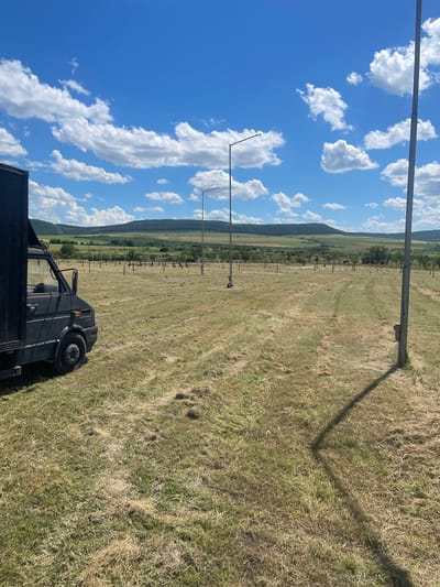 A dark panel van is parked on a grassy, partially mown field under a blue sky with scattered white clouds. The van is positioned on the left side of the frame, its rear facing towards the camera. In the background, a landscape of rolling hills and fields stretches out, with trees dotted across the terrain. Several tall, slender lampposts are visible, spaced out in the field, suggesting it might be a park or an open public space. The ground is covered in dry grass and some patches of green. The overall impression is of a rural or semi-rural setting on a bright, sunny day.