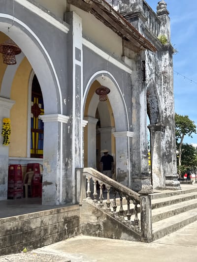 This image depicts the exterior of an old, weathered building, likely a religious or historical site, in Hội Tín, Vietnam. The architecture features crumbling concrete and plaster, with visible signs of age and decay. Arched doorways and windows, some with stained glass, are prominent.

The scene appears to be set during the daytime, with bright sunlight casting shadows. The sky is a clear blue. The building's facade is painted in a combination of gray and white, with some areas showing peeling paint and water stains. Decorative elements, such as ornate railings and decorative molding, are also present, though many are damaged.

In the foreground, several red plastic chairs are stacked, suggesting a temporary setup or that the area is used for gatherings. A woven basket-like object hangs from one of the arches, adding a rustic touch. In the background, lush green trees are visible, contrasting with the aged structure.

A single person, wearing dark clothing, is partially visible inside one of the arched openings, perhaps an attendant or a visitor. The overall atmosphere is one of historical significance and serene neglect. There is no visible text in the image.