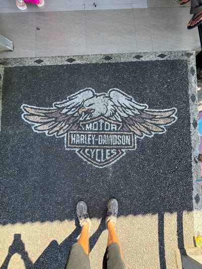 In Baturiti, Indonesia, a Harley-Davidson logo made of pebbles is embedded in a dark asphalt surface. The logo features an eagle with spread wings above a shield-shaped emblem containing the text "MOTOR HARLEY-DAVIDSON CYCLES". The logo is bordered by smaller pebbles and a concrete walkway. Two individuals are partially visible in the frame. One person, wearing flip-flops and pink clothing, stands on the walkway. Another person, wearing gray sneakers and shorts, stands on the asphalt, casting a shadow that stretches across the logo. The scene appears to be outdoors, under natural sunlight.