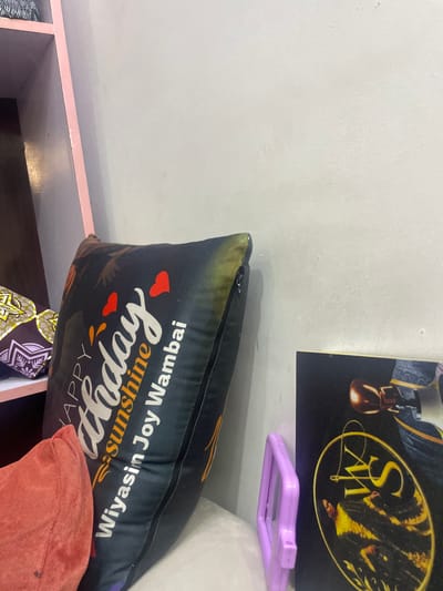 This image shows a pillow with the words "Happy Birthday Sunshine Wiyasin Joy Wambai" printed on it in white and yellow lettering. The pillow is black with an abstract design. There are also two small red hearts and some orange and yellow shapes on the pillow. The pillow is resting on a shelf, and a pink object is visible behind it. A framed picture is on the wall above the pillow. The picture is dark with a yellow oval logo in the center. The overall scene appears to be indoors, possibly in a home or a shop. The image is taken from a slightly tilted angle.