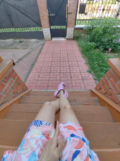 A first-person perspective shot shows a person sitting on wooden steps. The person is wearing pink Crocs and a colorful patterned skirt with blue, white, and orange leaf-like designs. The person's legs are crossed. The steps are weathered and show peeling brown paint. The background features a red brick wall, a red brick paved walkway, a black metal gate with decorative scrollwork, and green foliage. The location is identified as Krestovo-Gorodishche, Russia.