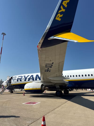 This media file captures a side view of a Ryanair airplane parked on an airport tarmac under a clear, bright blue sky, suggesting daytime with sunny weather. The image is oriented vertically. The plane is predominantly white with blue and yellow accents. Prominently visible on the tail is the word "RYANAIR" in large yellow letters against a dark blue background. The tail itself features a distinctive yellow winglet.

On the fuselage, the registration number "700-H6" is clearly marked in dark blue lettering on a silver section of the aircraft. The word "RYANAIR" is also emblazoned along the side of the plane in large, white, stylized lettering outlined in blue, with a design element resembling bird wings. Passengers are seen disembarking or boarding the aircraft via a set of portable stairs positioned at the rear exit.

In the foreground and background, tarmac surfaces are visible, marked with various painted lines and symbols, typical of an airport environment. Two orange and white traffic cones are placed on the ground near the aircraft. In the distance, a red and white striped pole with lights, possibly an aviation obstruction light, can be seen. The overall scene conveys a typical operational day at an airport. The image was taken in Fiumicino, Italy.
