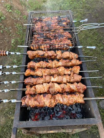 The image shows a close-up, overhead view of a barbecue grill in what appears to be an outdoor setting. The grill is filled with charcoal that is glowing red hot. On top of the grill, there are multiple skewers of meat being cooked. In the foreground, there are five skewers of cubed meat, each piece browned and slightly charred from the heat. Above these, there is a metal grilling basket containing what looks like chicken pieces, also showing signs of being cooked. Wisps of smoke are rising from the grilling food. The background consists of green grass and dirt, suggesting a natural or park-like environment. The overall scene evokes a feeling of outdoor cooking and grilling.