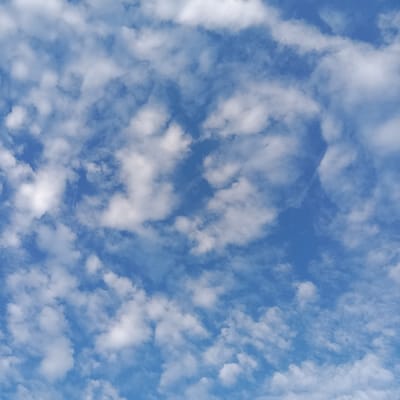 A clear blue sky is sparsely dotted with small, puffy white clouds. The clouds are distributed unevenly, with some areas showing more concentrated clusters and other areas revealing the unobstructed blue of the sky. The cloud formations are soft and rounded, creating a textured yet serene atmosphere.