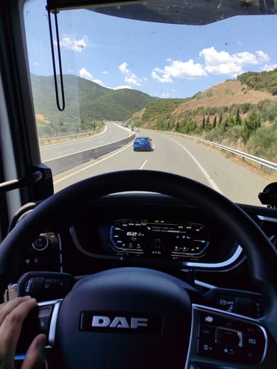 The image shows the view from the driver's seat of a DAF truck traveling on a highway in the Strymoniko Municipal Unit, Greece. The dashboard indicates a speed of 62 km/h. A blue car is visible in the lane ahead. The landscape consists of mountains covered with green vegetation under a blue sky with scattered clouds. The road has multiple lanes and is flanked by guardrails. The driver's hand is visible near the steering wheel.