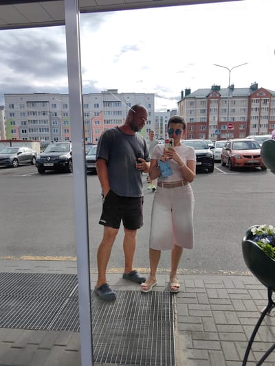 This media file captures a reflection in what appears to be a large window or mirror, showing two people standing in an outdoor parking lot. The setting is Fanipal, Belarus, indicated by the presence of apartment buildings with distinct architectural styles commonly found in post-Soviet countries and a sign with Cyrillic text.
On the left, a man wearing a grey t-shirt and black shorts is holding a phone and looking towards the right. He is wearing dark blue crocs. On the right, a woman with short hair and sunglasses is holding up her phone to take a selfie. She is wearing a light pink t-shirt, light pink culottes, and beige sandals. A small blue object, possibly a pouch or bag, is hanging from her hand.
The scene is a parking lot with several cars visible in the background, including a black Renault. The sky is overcast with a mix of light and dark clouds, suggesting the time of day is likely afternoon. The overall mood appears casual, with the couple engaged in taking a photograph of themselves. The reflection shows them standing on a paved area with a metal grate in the foreground. The apartment buildings in the background are multi-story structures with rows of windows, painted in various pastel colors. There is no visible text in the image beyond the license plates of the cars, which are not clearly legible.
